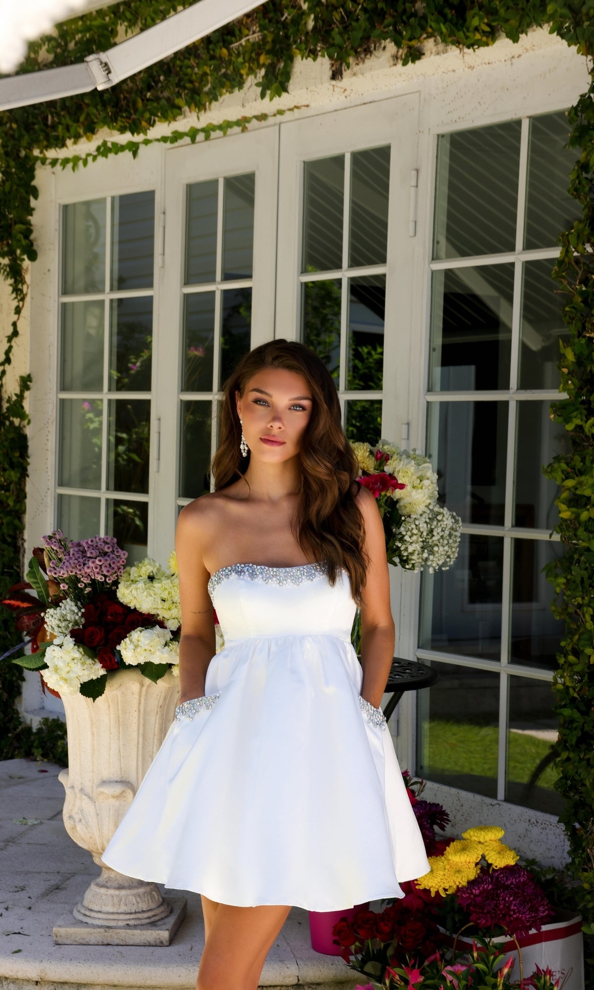 A woman wearing the Strapless Short Hoco Dress: Ava Presley 29837 stands by large windows, surrounded by potted flowers and lush greenery.